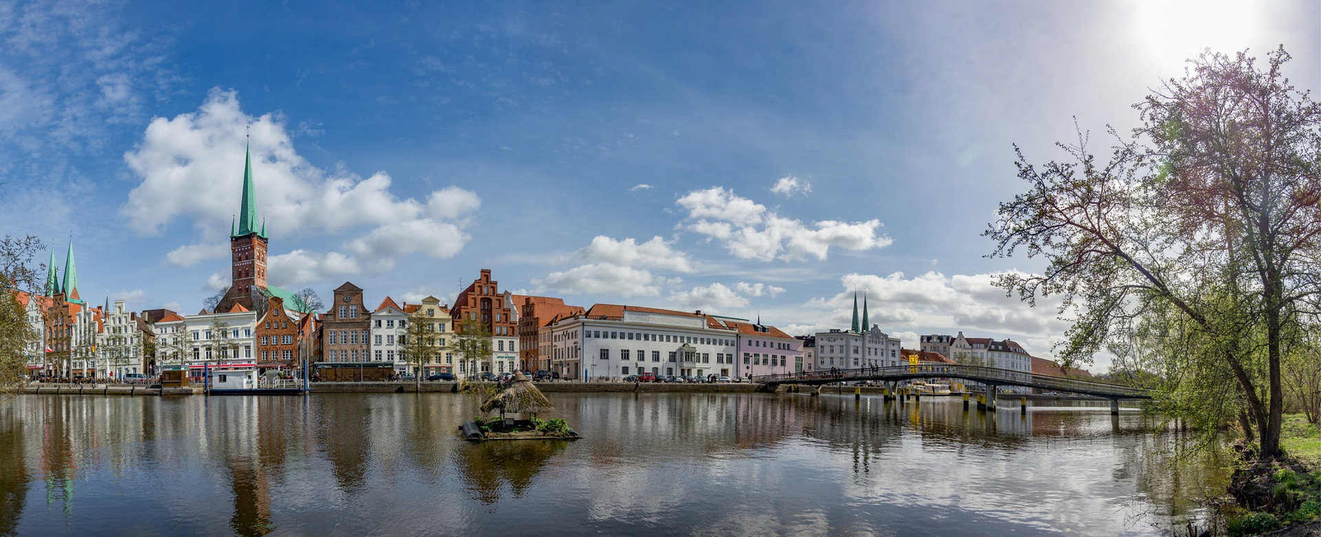Ansicht der Lübecker Altstadt im Bereich der Obertrave. Im Vordergrund der Fluß, dahinter die Straße mit stattlichen Kaufmannshäusern und der Musikschule. Über den Fluß führt eine Brücke zur Hochschule. Im linken Bereich sind Dach und Turm der Petrikirche sichtbar, rechts noch die beiden Türme des Doms.
