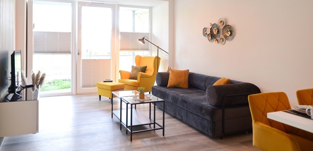 Bild einer Gästewohnung der TRAVE mit gemütlichem Sofa und einem Ohrensessel. Links sieht man einen Flychbildfernseher, rechts eine Essgruppe. das gemütliche Sofa, der Couchtisch und ein Ohrensessel mit Fußhocker