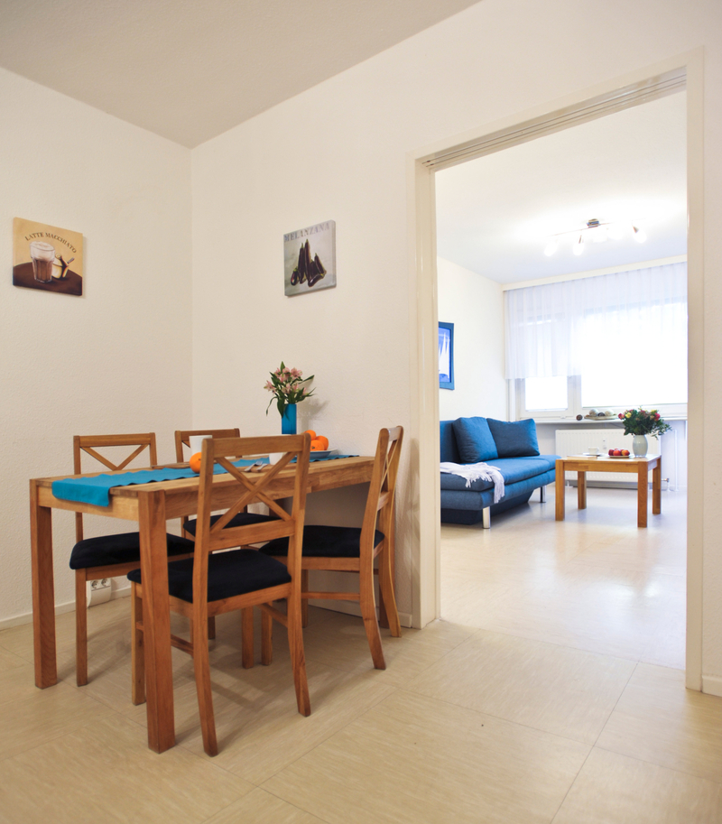 Vorn im Bild der Esstisch und vier Stühle, im Hintergrund durch einen Durchgang sichtbar, die Couch mit dem Couchtisch vor dem Fenster