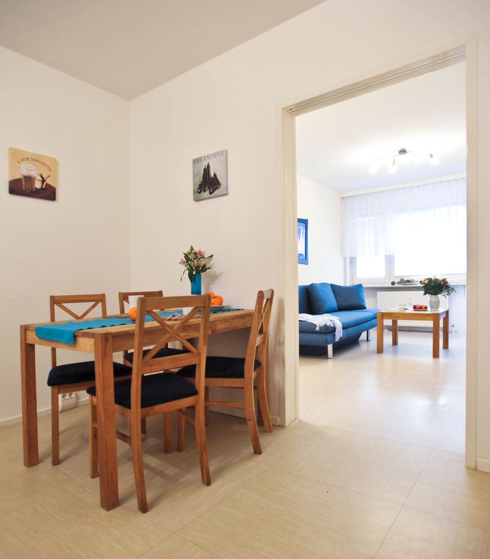 Vorn im Bild der Esstisch und vier Stühle, im Hintergrund durch einen Durchgang sichtbar, die Couch mit dem Couchtisch vor dem Fenster
