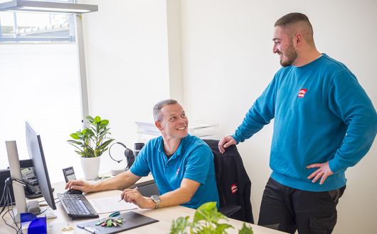 Zwei Hauswarte der TRAVE. Einer sitzt am Schreibtisch vor dem Computer, der andere steht hinter im. Beide schauen sich an und lächeln.