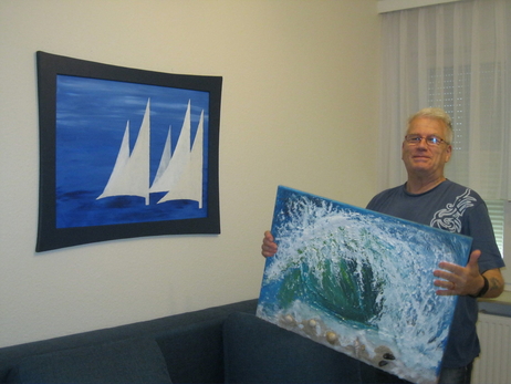 Ein ehrenamtlich Tätiger hält ein selbstgemaltes Bild in der Hand. An der Wand dahinter befindet sich ein weiteres Bild, das im TRAVE-Treff entstanden ist.