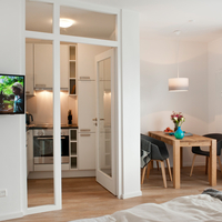 Blick in den Raum: Flachbildfernseher an der Wand, rechts Esstisch mit zwei Stühlen, dazischen der Blick in die Küche.