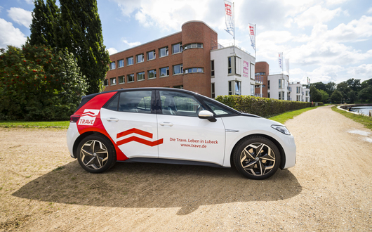 Ein Elektroauto mit dem TRAVE-Design vor dem Gebäude der TRAVE auf der Seite der Kanal-Trave (Fluß).
